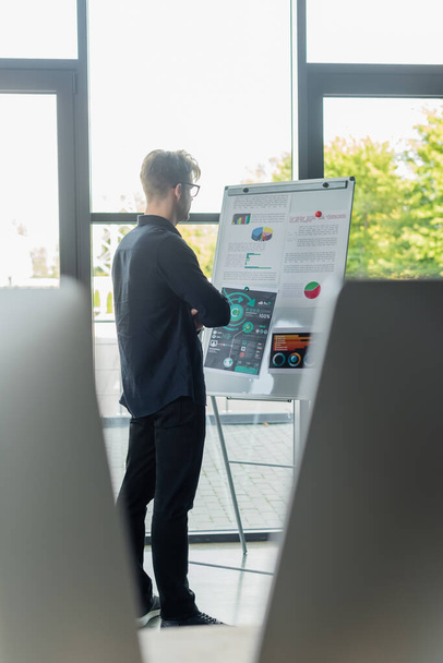 Programátor prohlíží grafy na flip grafu poblíž rozmazaných monitorů počítače v kanceláři  - Fotografie, Obrázek