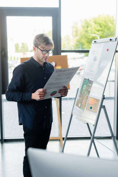Programmer in eyeglasses holding document near flip chart and blurred computer in office  - Foto, Bild