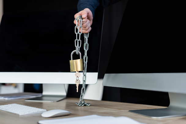 Cropped view of programmer holding padlock on chain near computers in office  - Photo, Image
