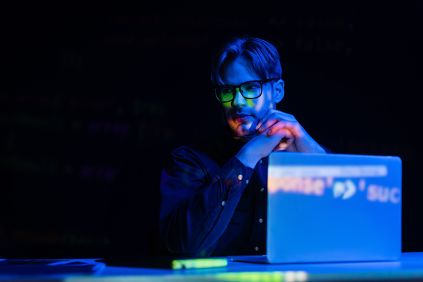 Programista w okularach siedzi w pobliżu komputera z odbiciem kodu w biurze odizolowany na czarno  - Zdjęcie, obraz