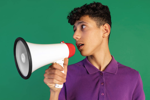 Cropped portrait of young boy in purple T-shirt speaking into megaphone isolated over green background. Giving information - Photo, Image