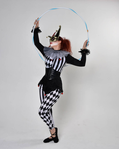 Full length  portrait of red haired  girl wearing a black and white clown jester costume, theatrical circus character.  Standing pose with hula hoop, isolated on  studio background. - Photo, Image