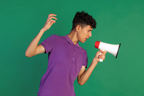 Cropped portrait of young boy in purple T-shirt talking in megaphone isolated over green background. Sharing information - Photo, Image
