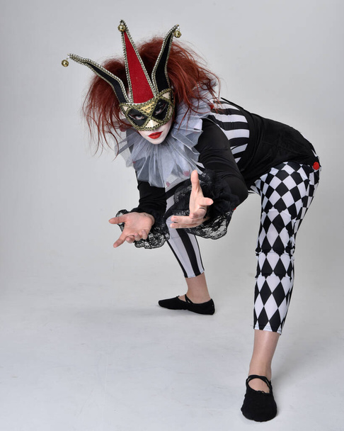 Portrait complet de fille aux cheveux roux portant un costume de bouffon de clown noir et blanc, personnage de cirque théâtral. Assis sur le sol, isolé sur le fond du studio. - Photo, image