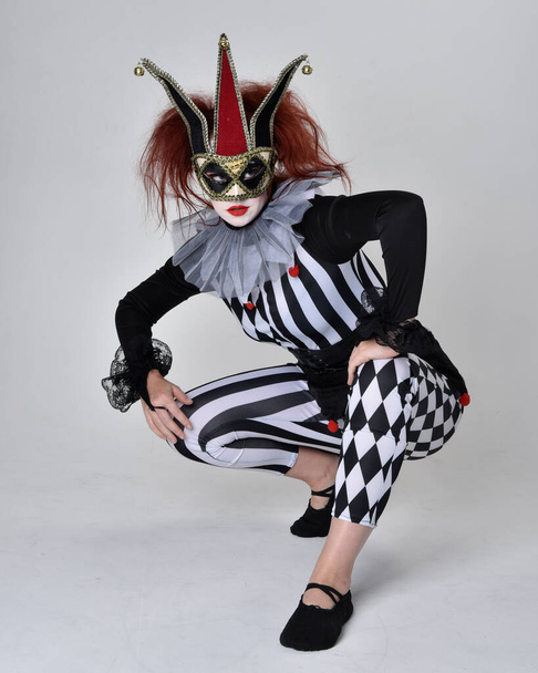 Полнометражный портрет рыжеволосой девушки в чёрно-белом костюме шута-клоуна, театрального циркового персонажа. Сидя на полу, изолированный на студийном фоне. - Фото, изображение