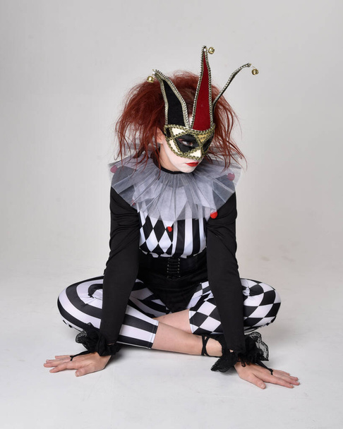 Retrato de comprimento total da menina de cabelos vermelhos vestindo um traje de bobo de palhaço preto e branco, personagem de circo teatral. Sentado no chão, isolado no fundo do estúdio. - Foto, Imagem