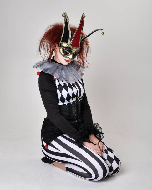 Full length  portrait of red haired  girl wearing a black and white clown jester costume, theatrical circus character.  Sitting down on floor, isolated on  studio background. - Photo, Image