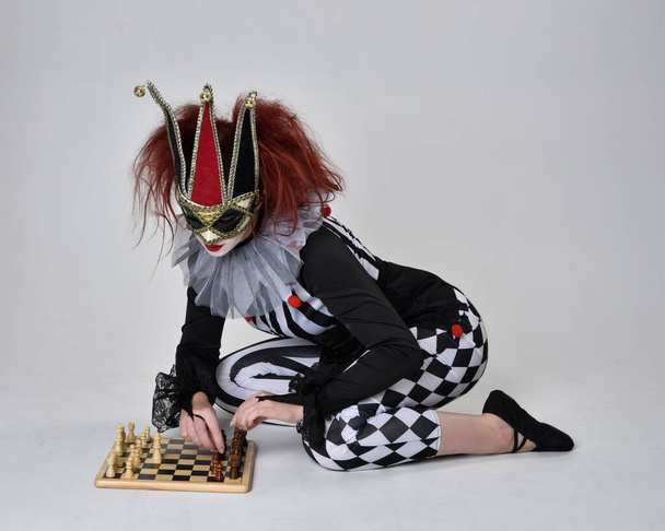 Volledig portret van een roodharig meisje in een zwart-wit clown nar kostuum, theatraal circusfiguur. Zittend op de vloer, geïsoleerd op studio-achtergrond. - Foto, afbeelding