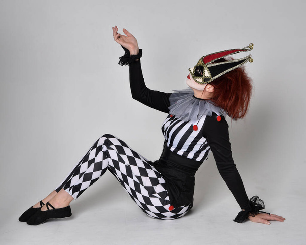 Full length  portrait of red haired  girl wearing a black and white clown jester costume, theatrical circus character.  Sitting down on floor, isolated on  studio background. - Foto, Bild
