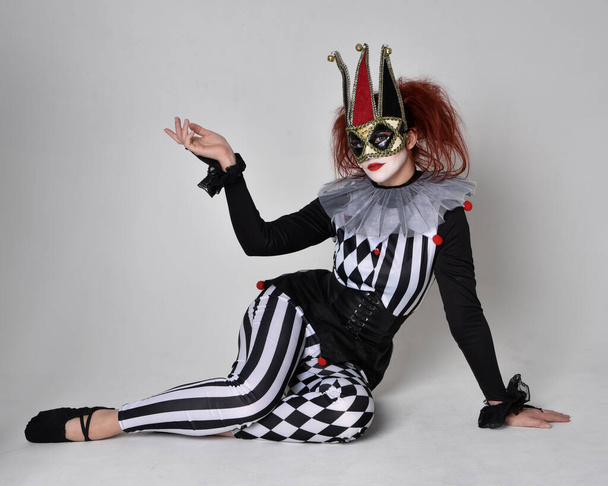 Portret rudej dziewczyny w czarno-białym kostiumie błazna klauna, teatralny charakter cyrku. Siedząc na podłodze, odizolowany na tle studia. - Zdjęcie, obraz