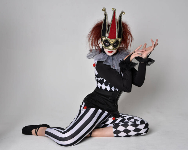 Ganztägiges Porträt eines rothaarigen Mädchens, das ein schwarz-weißes Clownskostüm trägt, eine theatralische Zirkusfigur. Auf dem Boden sitzend, isoliert auf Studiohintergrund. - Foto, Bild