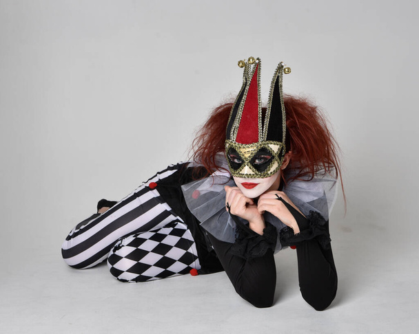 Ganztägiges Porträt eines rothaarigen Mädchens, das ein schwarz-weißes Clownskostüm trägt, eine theatralische Zirkusfigur. Auf dem Boden sitzend, isoliert auf Studiohintergrund. - Foto, Bild