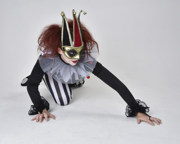 Dlouhý portrét rudovlasé dívky v černobílém kostýmu klauna šaška, divadelní postava cirkusu. Sedí na patře, izolovaný na pozadí studia. - Fotografie, Obrázek