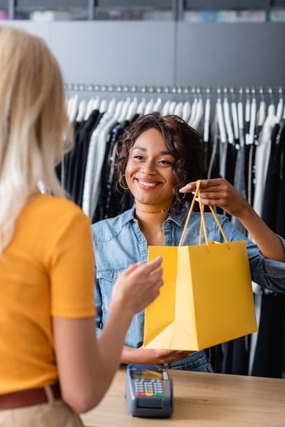 smiling african american saleswoman giving yellow paper bag with purchase to blonde client in boutique - Photo, Image