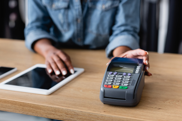 cropped view of african american saleswoman holding payment terminal near gadgets on sales counter desk - 写真・画像