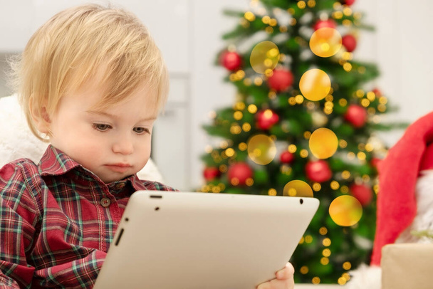 Caucasian baby girl of 1-2 year looking at tablet indoors.Concept of child watching cartoons,Christmas film,playing computer game or reading electronic book.Toddler with gadget on New Year holidays. - Photo, image