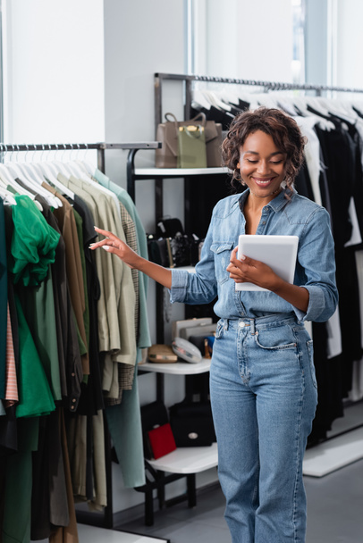 fröhliche afrikanisch-amerikanische Verkäuferin hält digitales Tablet in der Nähe von Kleidung auf dem Regal - Foto, Bild