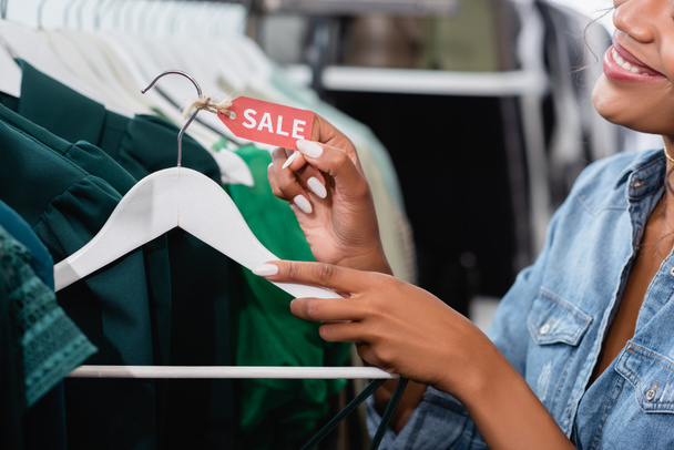 vue recadrée de joyeux vendeur afro-américain tenant cintre avec étiquette de vente dans la boutique de vêtements - Photo, image