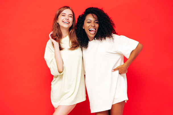 Two young beautiful smiling international hipster female in trendy summer clothes. Sexy carefree women posing near red wall in studio. Positive models having fun. Concept of friendship - Photo, image