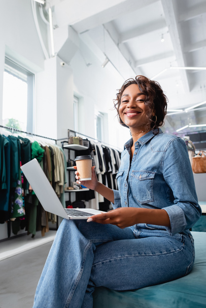 happy african american woman holding laptop and paper cup in clothing boutique  - Foto, immagini