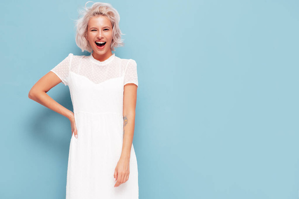 Портрет молодой красивой улыбающейся женщины в модном летнем белом платье. Сексуальная беззаботная блондинка позирует возле синей стены в студии. Позитивная модель развлекается в помещении. Весёлые и счастливые - Фото, изображение