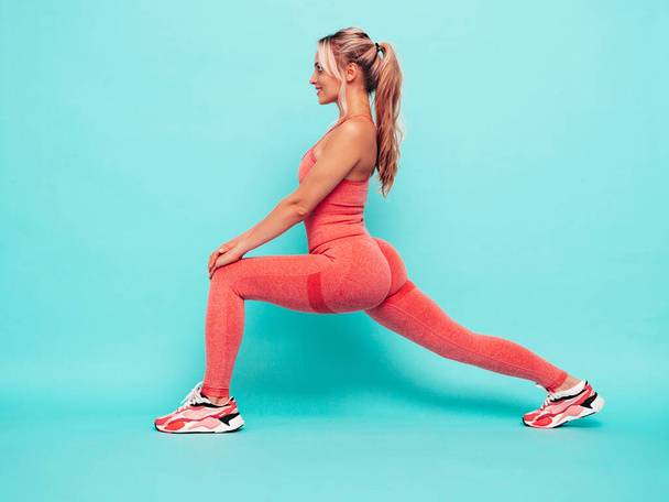 Fitness smiling woman in pink sports clothing. Young beautiful model with perfect body.Female posing near blue wall in studio.Cheerful and happy. Stretching out before training. Doing lunges - Foto, imagen