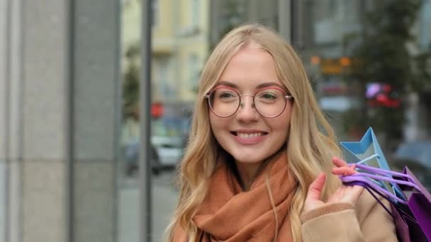 Portret Kaukasische vrolijke vrouw vrouwelijke shopper glimlachen houden in de hand heldere geschenkzakjes jonge dame in glazen staan op straat en wachten op vriend meisje genieten van kortingen en verkopen in het centrum van de stad - Video