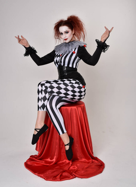 Ganztägiges Porträt eines rothaarigen Mädchens, das ein schwarz-weißes Clownskostüm trägt, eine theatralische Zirkusfigur. sitzende Pose, isoliert auf Studiohintergrund. - Foto, Bild