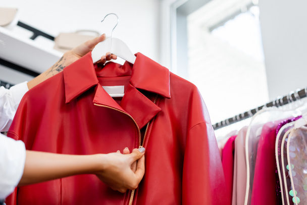 przycięty widok wytatuowanej kobiety trzymającej czerwoną skórzaną kurtkę na wieszaku przy wyborze ubrania  - Zdjęcie, obraz