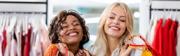 alegres mujeres interracial sosteniendo bolsas de compras con compras en boutique de ropa, pancarta  - Foto, imagen
