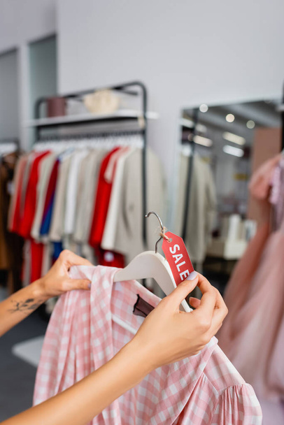 partial view of tattooed young woman holding hanger with checkered pink dress and sale tag  - Foto, immagini