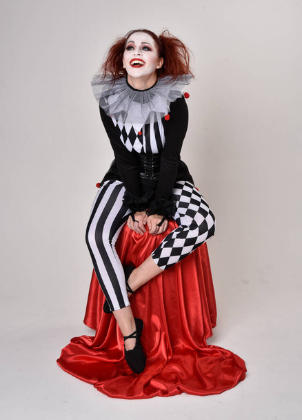 Portret rudej dziewczyny w czarno-białym kostiumie błazna klauna, teatralny charakter cyrku. pozycja siedząca, odizolowana na tle studia. - Zdjęcie, obraz