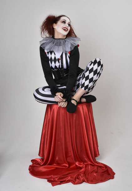 Dlouhý portrét rudovlasé dívky v černobílém kostýmu klauna šaška, divadelní postava cirkusu. sedící póza, izolované na pozadí studia. - Fotografie, Obrázek