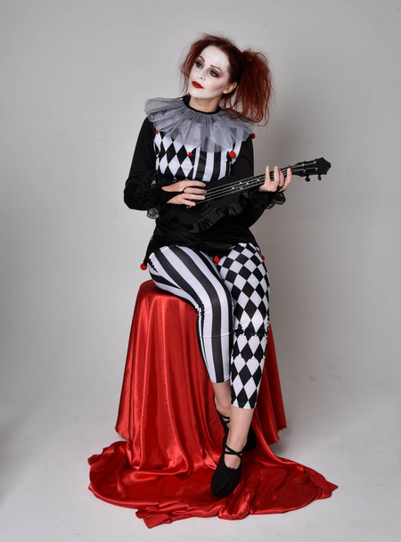 Full length  portrait of red haired  girl wearing a black and white clown jester costume, theatrical circus character.    sitting pose, isolated on  studio background. - Foto, imagen