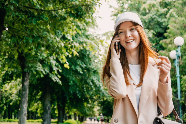 Zrzavá žena s venkovním portrétem na mobilu. Happy Young obchodní žena s mobilním telefonem v ruce procházky zeleným letním parkem - Fotografie, Obrázek