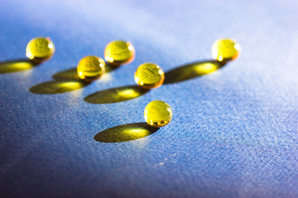 Yellow colored fish oil pills, tablets, omega 3 gelatin capsules lie on blue table leaving contrasting long shadows flatly. Medical, health care, medicine treatment minimalistic background top view. - Photo, Image