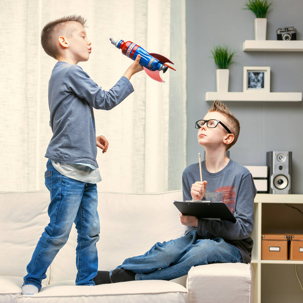 Netter kleiner Junge spielt mit einer Spielzeugrakete auf einem Sofa, beobachtet von seinem Bruder, der neben ihm sitzt und an einem Klemmbrett arbeitet - Foto, Bild