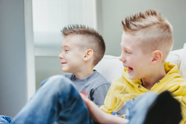 Lindo joven con un moderno peinado puntiagudo sentado en un sofá con su hermano riéndose mientras ven o juegan juegos en casa - Foto, imagen
