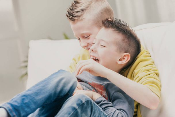 Δύο αγαπημένοι νεαροί αδερφοί απολαμβάνουν ένα τραχύ και κυλιόμενο στον καναπέ γελώντας χαρούμενα καθώς γαργαλιούνται σε μια στενή αγκαλιά - Φωτογραφία, εικόνα