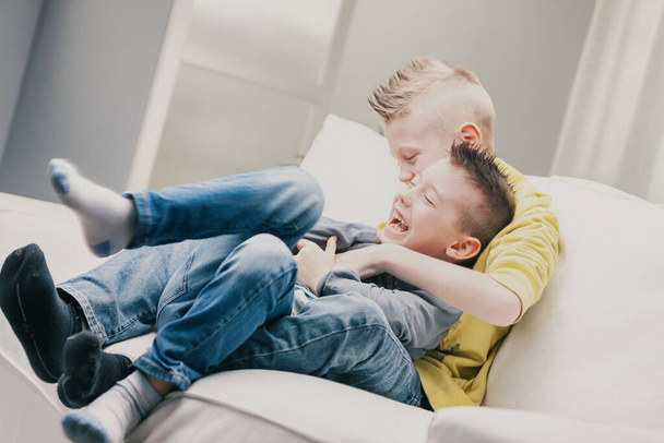 Два смеющихся маленьких мальчика играют на диване дома, обнимаются и щекочут друг друга, наслаждаясь проведением хорошего семейного времени вместе. - Фото, изображение