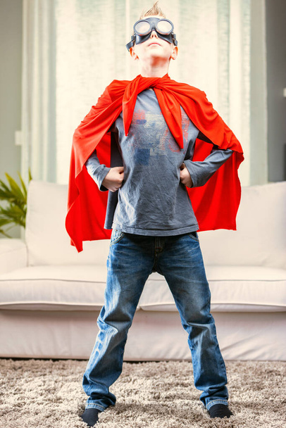 Stolzer junger Superheld posiert in rotem Umhang und Brille mit den Händen auf den Hüften im heimischen Wohnzimmer - Foto, Bild