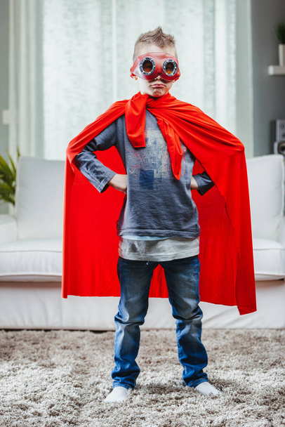 Zdeterminowany młody Super Hero w kolorowym czerwonym pelerynie stojący z rękami na biodrach i krzykiem wewnątrz domu - Zdjęcie, obraz