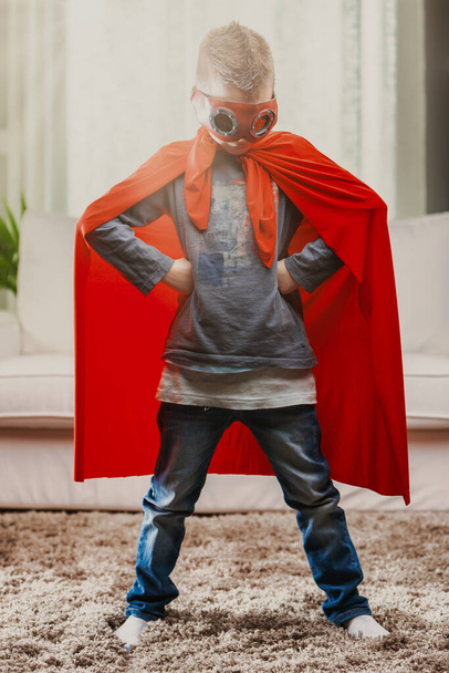 Young boy posing arms akimbo in a super hero costume with dramatic red cape indoors in the living room - Photo, Image