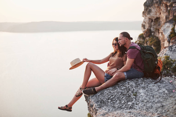Hugging couple with backpack sitting on top of rock mountain enjoying view coast a river or lake. Traveling mountains and coast, freedom and active lifestyle concept. - Fotografie, Obrázek