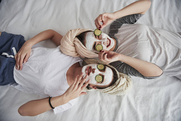 Taking off these things. Lying on the white bed. Top view. Conception of skin care by using white mask and cucumbers on the face. - Foto, afbeelding