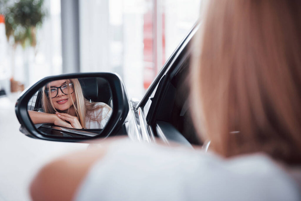 vrouw in bril kijkend in zijspiegel van een modern voertuig in de salon. - Foto, afbeelding