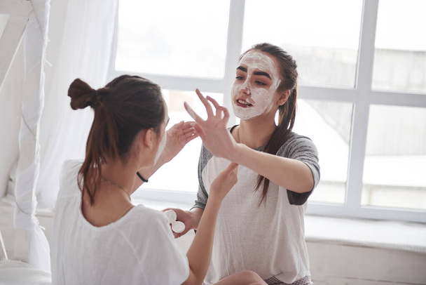 Sitting against white window. Conception of skin care by using white mask on the face. Two female sisters have weekend at bedroom. - Foto, immagini