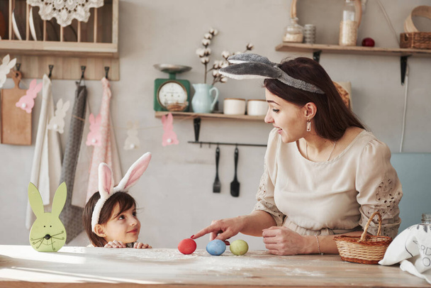 Frau zeigt rotes Ei mit Zeigefinger Mutter und Tochter in Hasenohren zu Ostern haben tagsüber Spaß in der Küche. - Foto, Bild