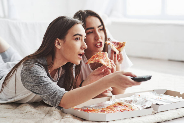 No way, did you saw that. Sisters eating pizza when watching TV while lying on the floor of beautiful bedroom at daytime. - Foto, immagini