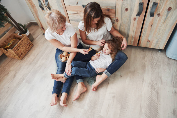 Kid lying on the parent's legs. Mother, grandmother and daughter having good time in the kitchen. - Foto, afbeelding